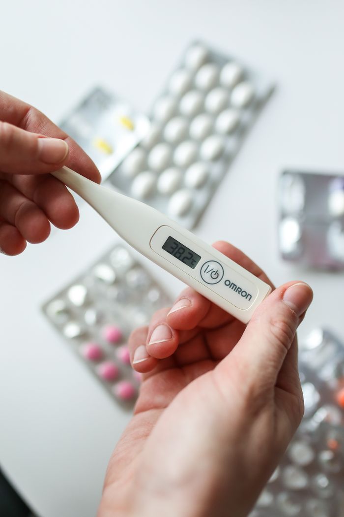 About Close-up of hands holding a digital thermometer with medication in the background.